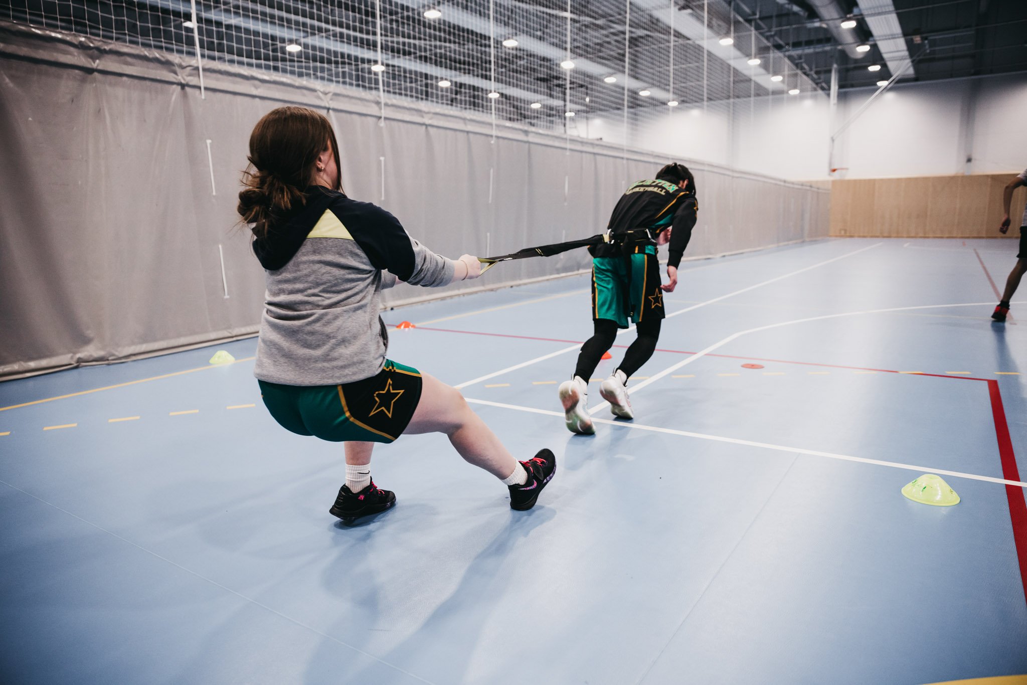To ungdommer forebygger skade i baskettrening. Jente holder igjen gutt som drar i et bånd innendørs i hall med blått gulv