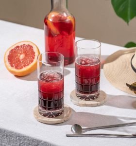 to glass med rød drikke og isbiter på et bord med hvit duk og vannkanne med rød drikke og oppskjært rød appelsin ved siden av med et planteblad i bakgrunnen. 