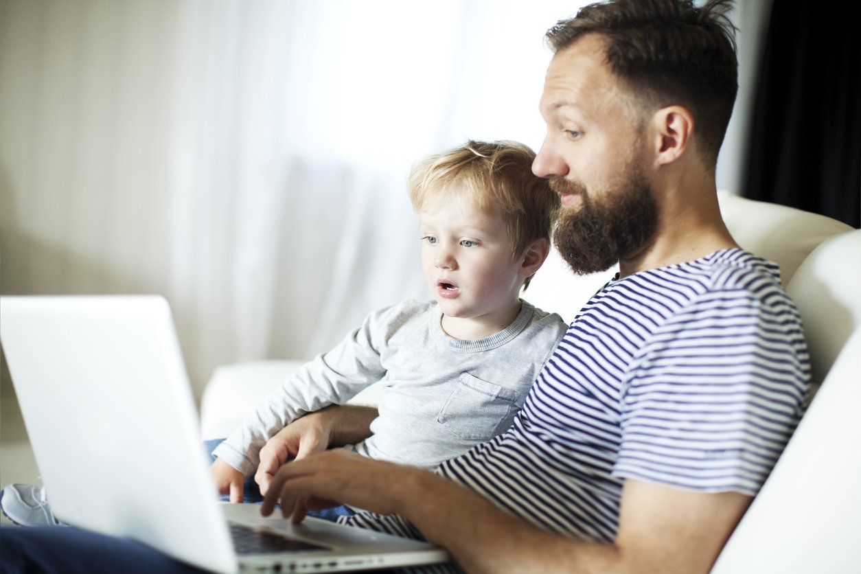 En far i hvit og blå stripete t-skjorte og sønn i en grå genser som sitter i en hvit sofa i stua og ser i en pc-skjerm sammen.