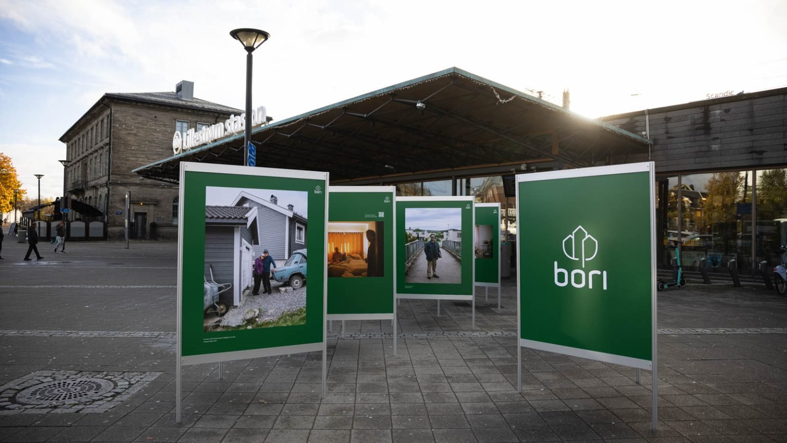 Bilde av grønne reklameplakater plassert på torget utenfor Lillestrøm Stasjon, med skyer og solen i bakgrunnen.