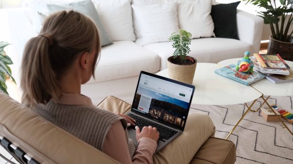 Kvinne som bruker en bærbar datamaskin på en sofa i en lys stue, med et salongbord og barneleker i bakgrunnen, inkludert en styreportal.