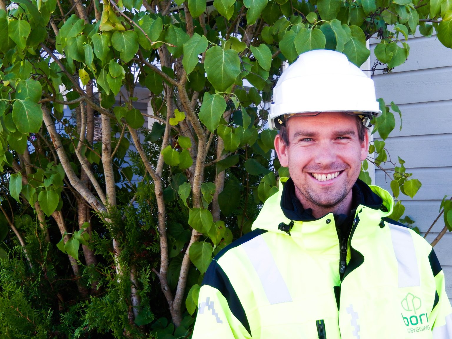 Stian smiler til kamera iført hvit hjelm og signalgul jakke under et besøk på en rehabiliteringen av et borettslag.