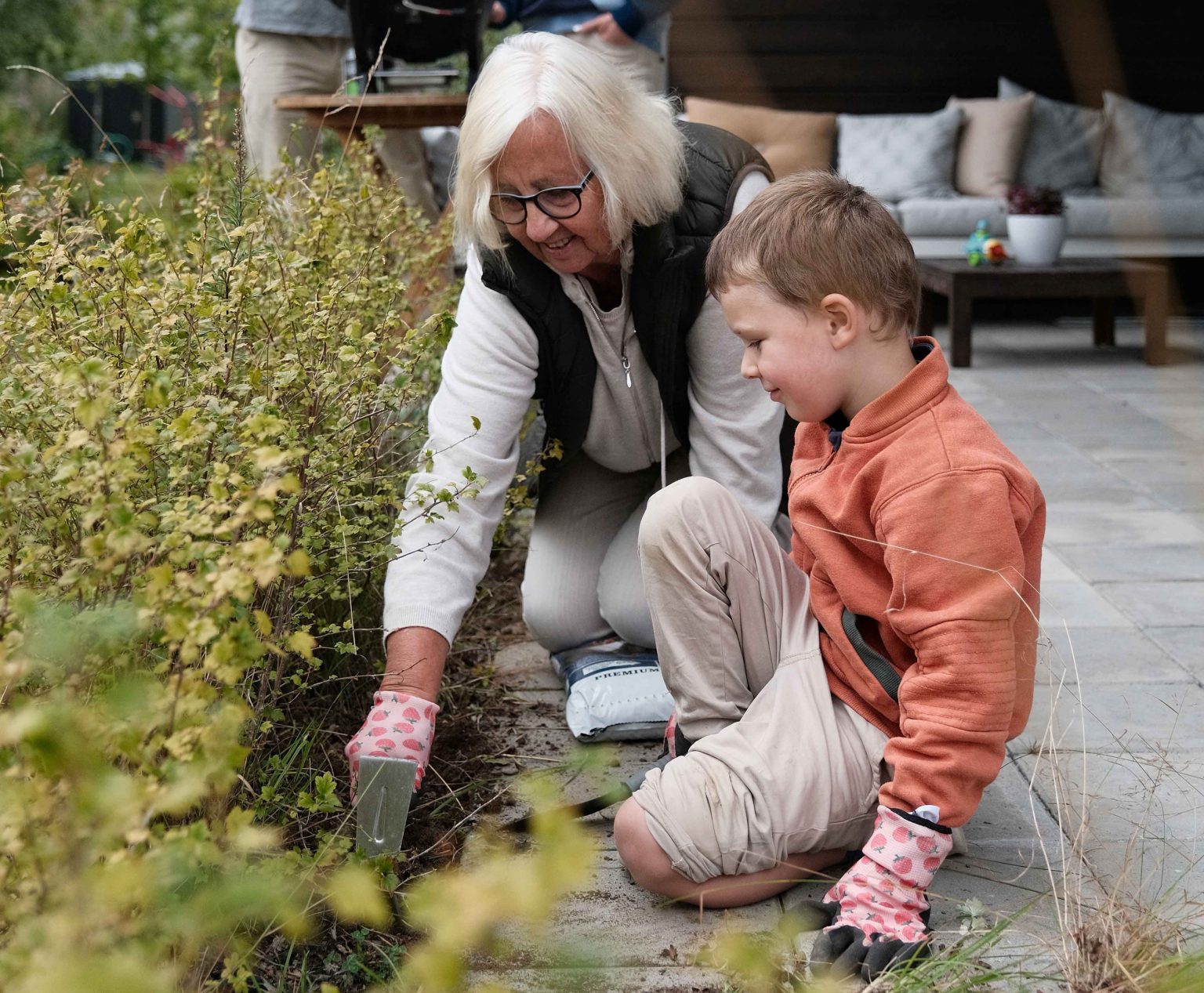 En eldre kvinne og en liten gutt sitter på huk i en hage. Kvinnen har på redskapshansker og viser gutten hagearbeid.