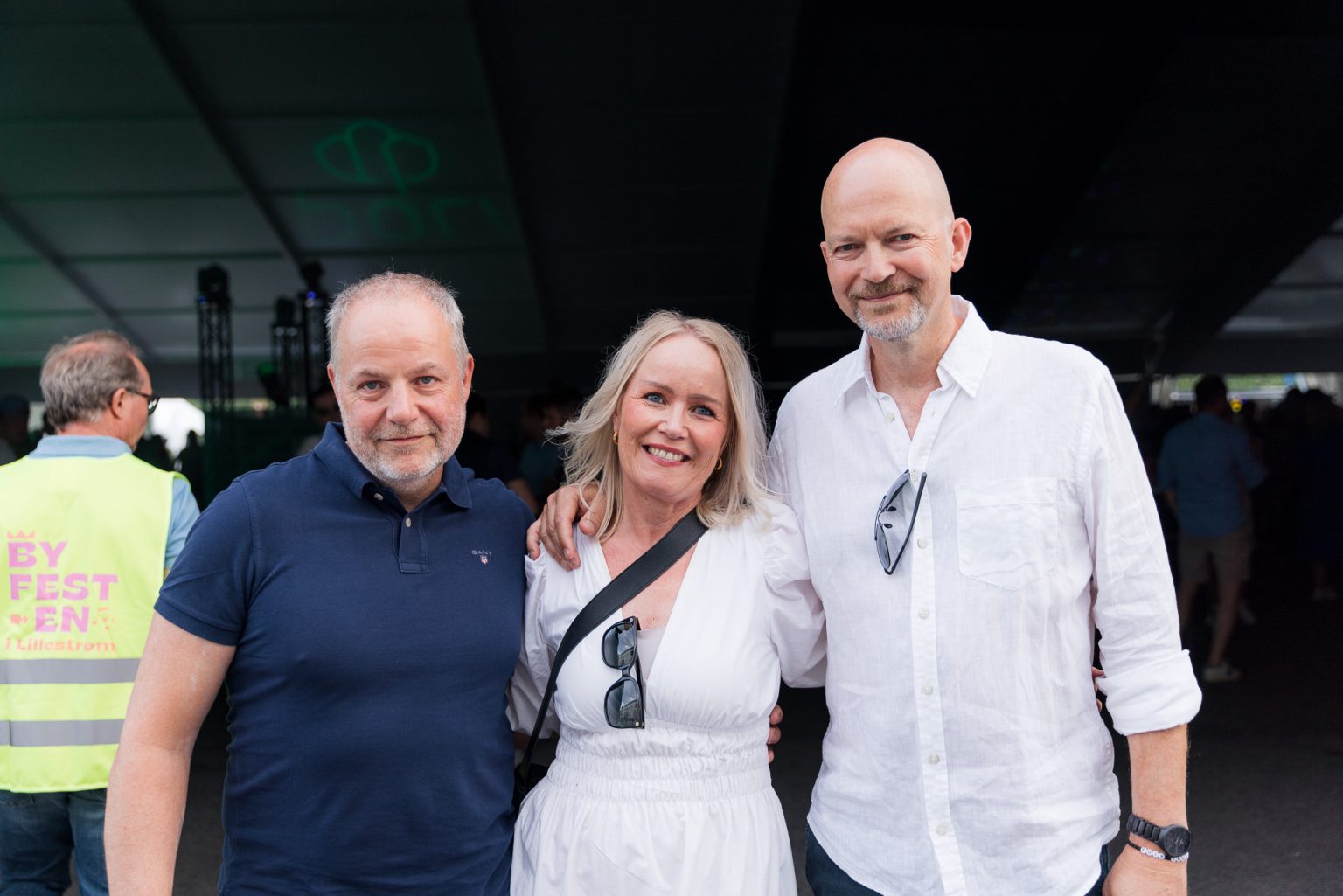 to menn i blå t-skjorte og hvit skjorte og en dame med hvit kjole som står sammen på konsertområdet og holder rundt hverandre og smiler inn i kameraet.