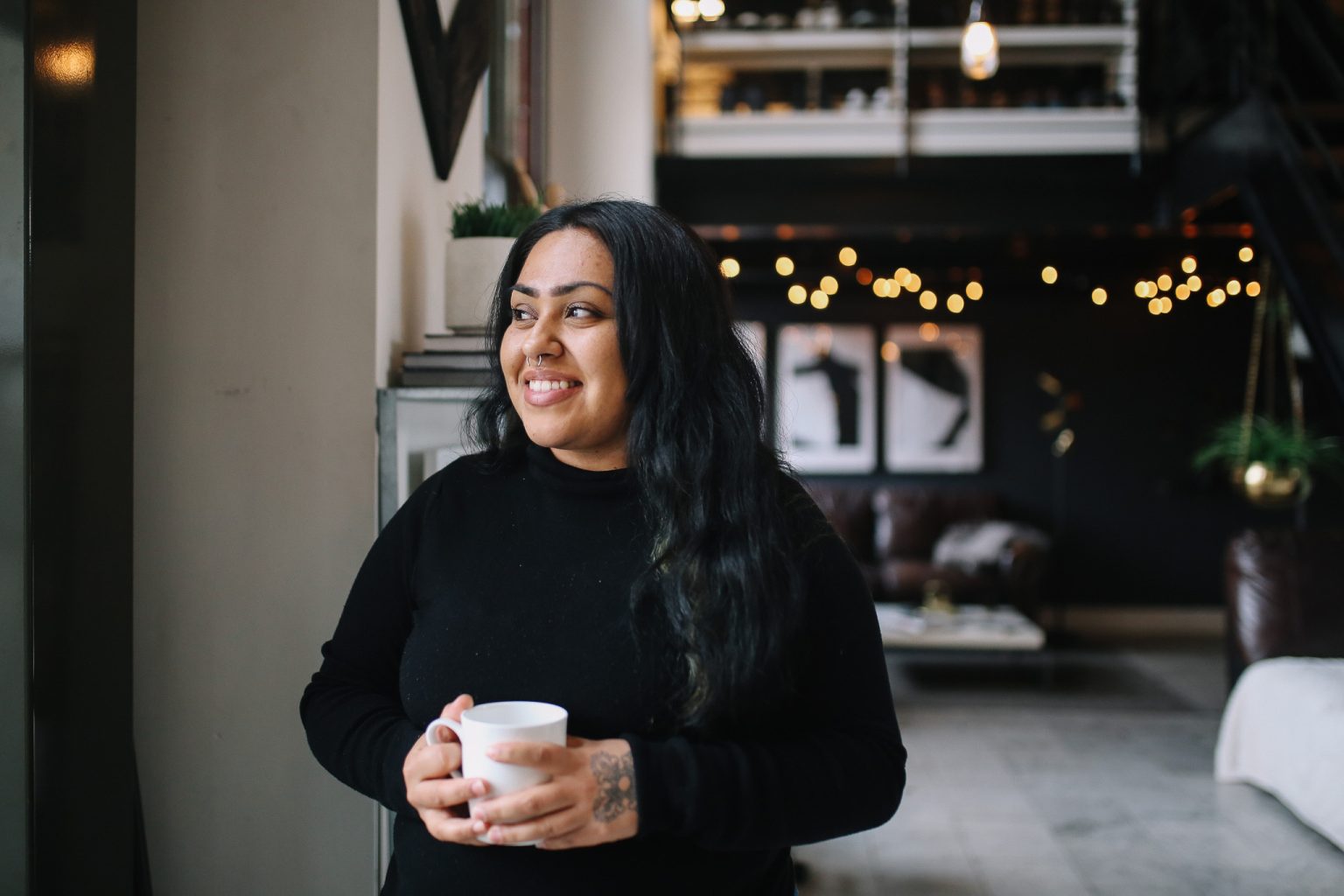 Ung mørk kvinne med langt sort hår, sorte klær og nesepiercing står smilende i en mørk stue med en hvit kaffekopp mellom hendene.