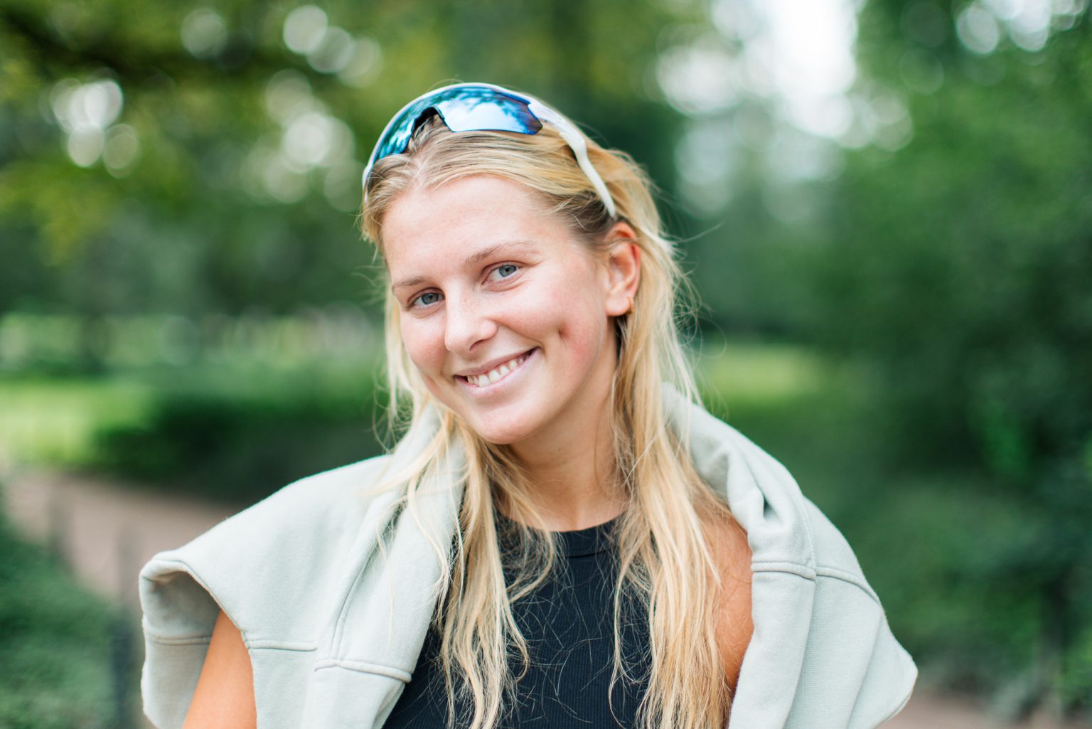 en dame med blondt langt hår, blå solbriller, sort topp og grå genser over skuldrene smiler inn i kameraet med grønne trær i bakgrunnen.