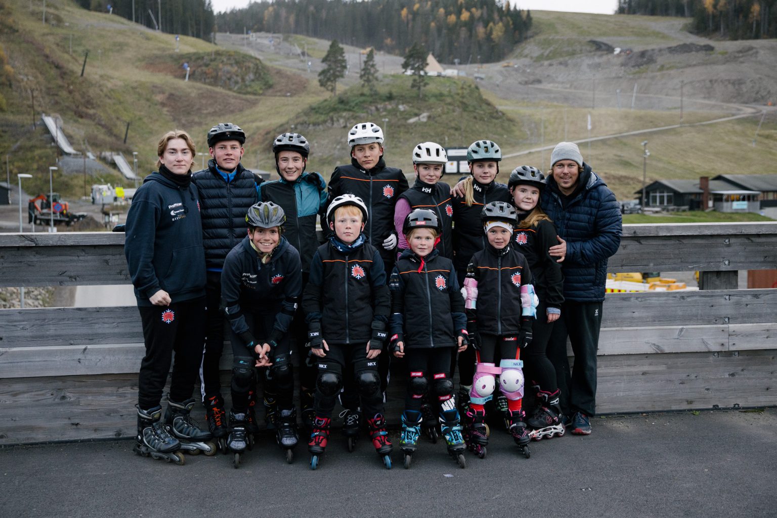 et idrettslag på 12 personer står på en bro foran en alpinbakke på høsten i treningsutstyr og smiler inn i kameraet