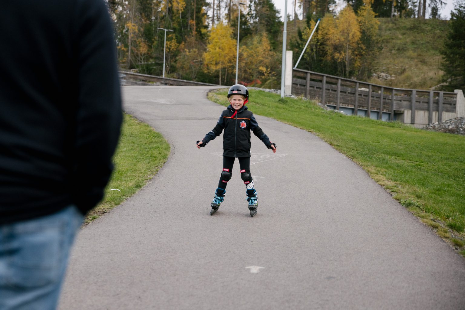 en ung jente i rollerbladesutstyr suser ned en asfaltbakke og smiler til en forelder som står i forgrunnen