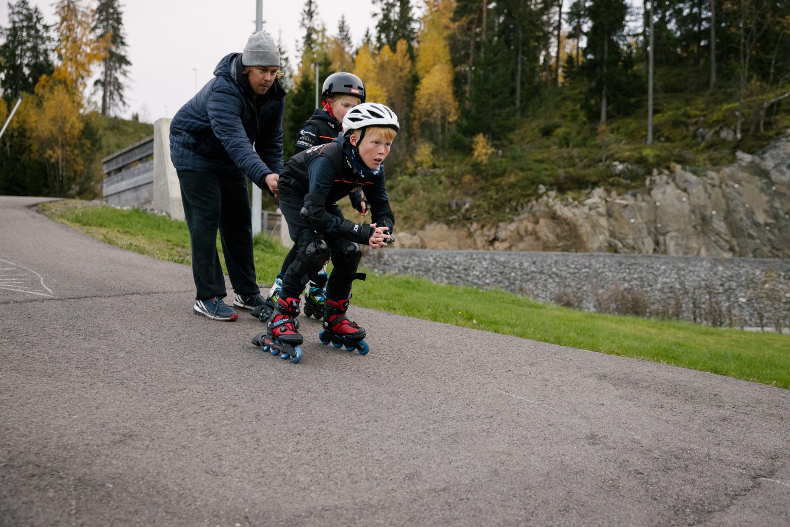 en gutt i rollerbladesutstyr er på vei ned en asfaltbakke med treneren som står i bakgrunnen