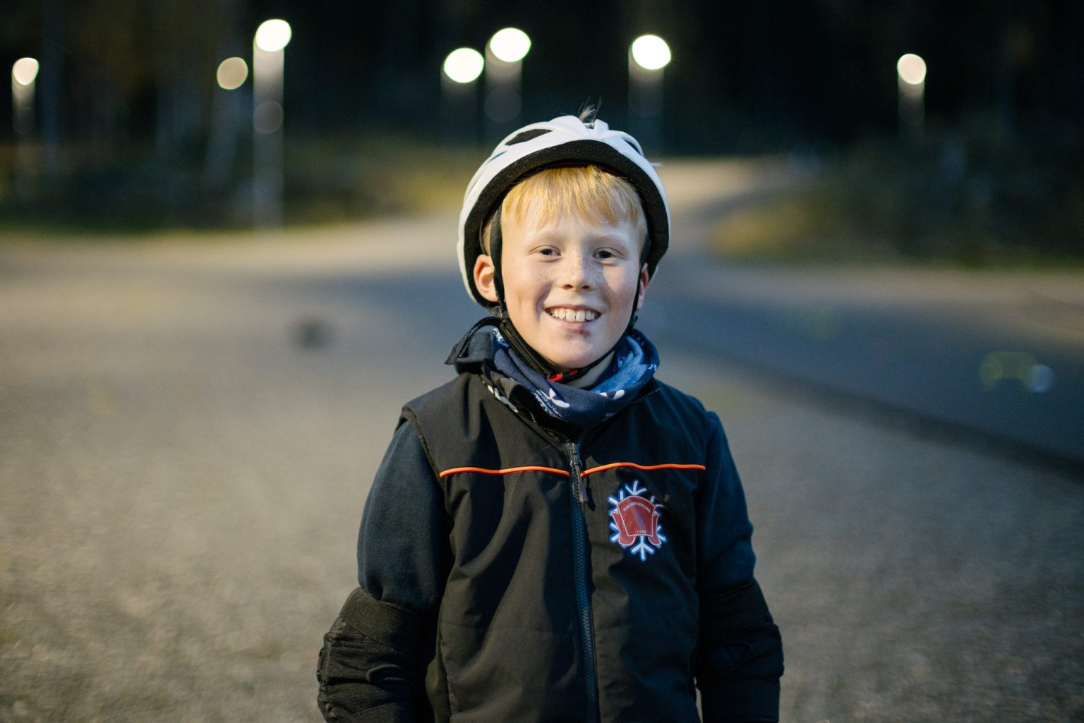 en gutt med hvit hjelm og sort treningsutstyr smiler inn i kameraet med skog og asfaltvei bak seg