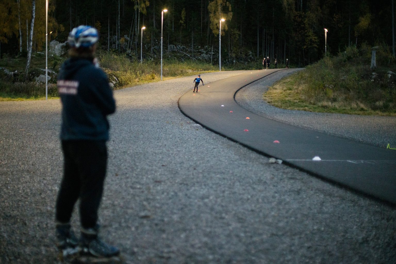en utøver suser ned en asfaltbakke med en trener i forgrunnen i høstmørket