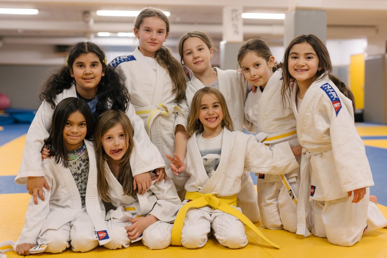en gjeng på åtte jenter iført hvite judodrakter med gule belter som sitter samlet i en treningssal og holder rundt hverandre og smiler inn i kameraet.