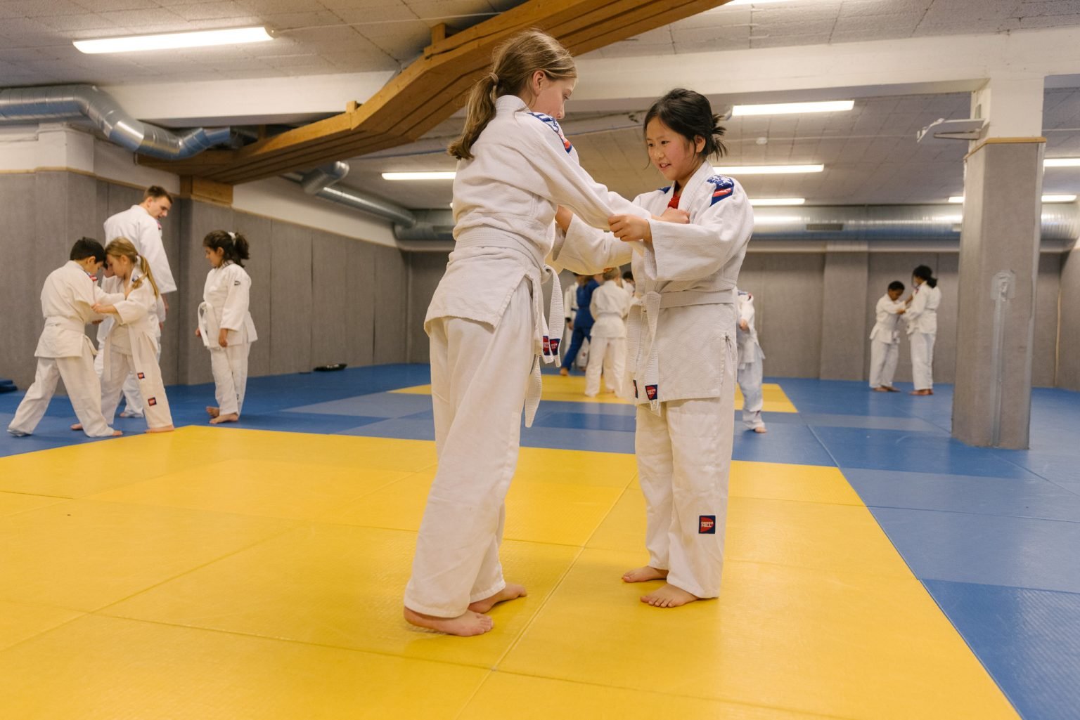 to jenter iført judodrakter holder i hverandre like før de skal utføre bakkekampøvelser i en treningssal med gulvet dekket av madrasser i blått og gult. I bakgrunnen sees andre idrettsutøvere som også trener.