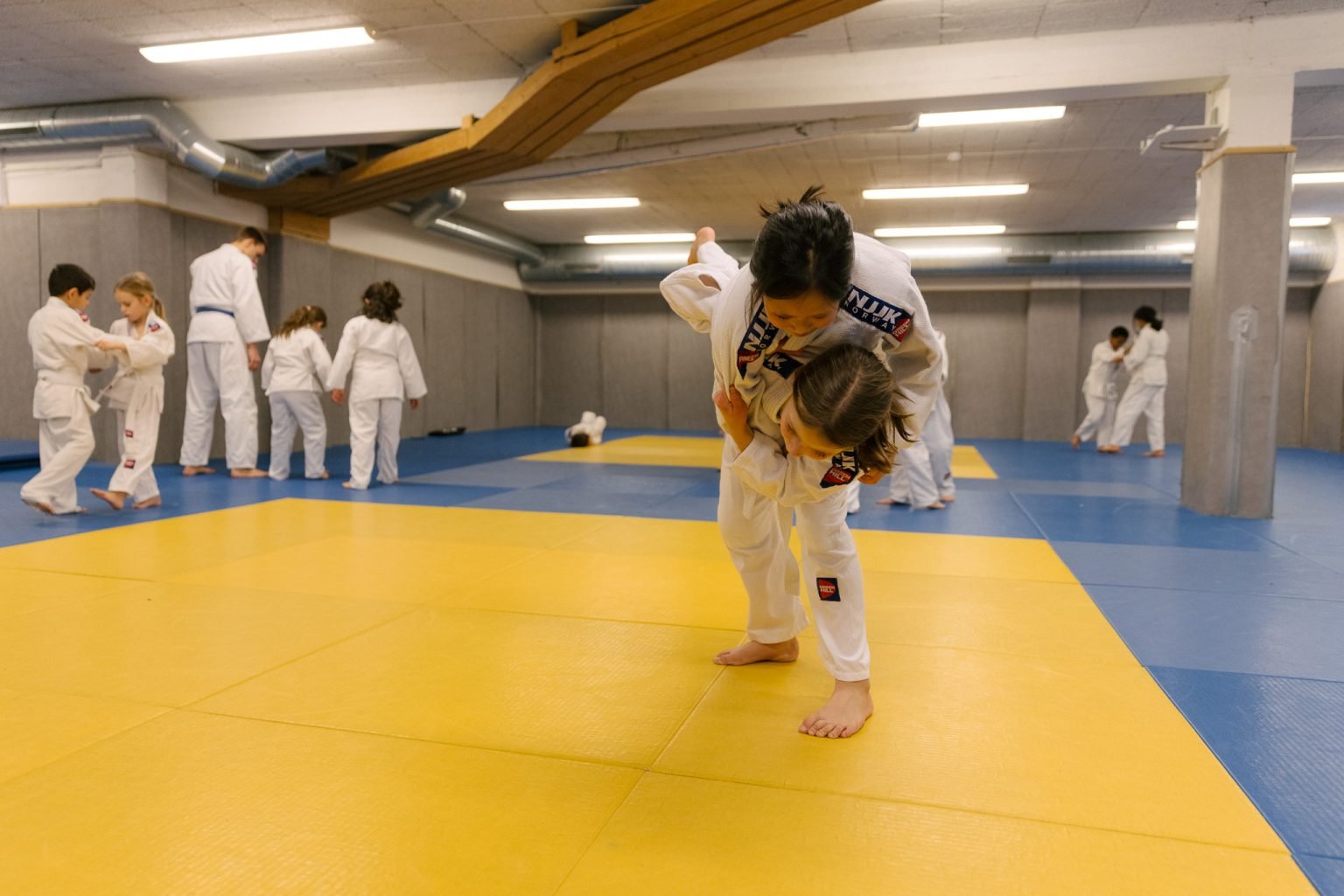 to jenter iført judodrakter utfører bakkekampøvelser i en treningssal med gulvet dekket av madrasser i blått og gult. I bakgrunnen sees andre idrettsutøvere som også trener.