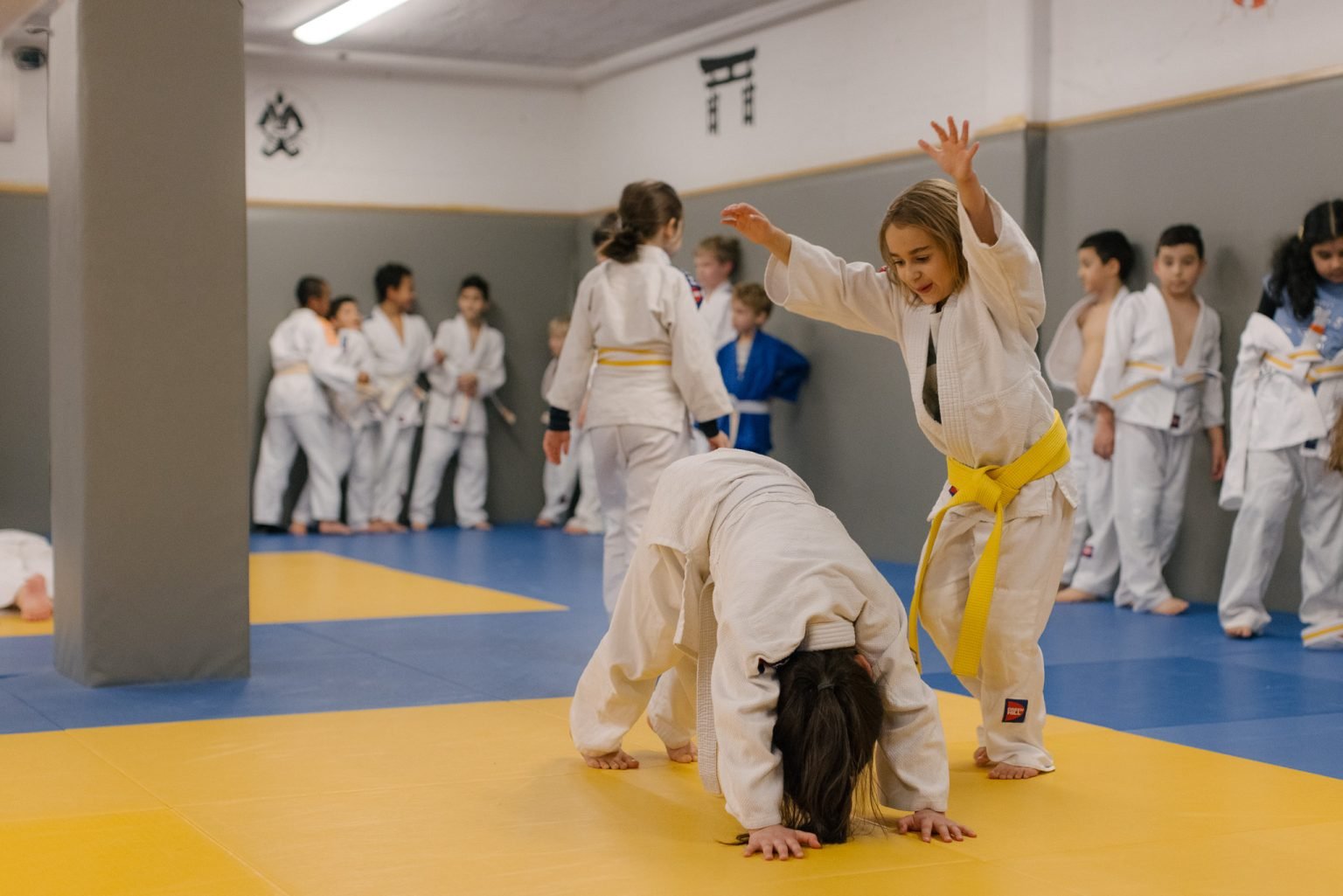 en jente iført hvit judodrakt med gult belte hopper bukk over en annen utøver.