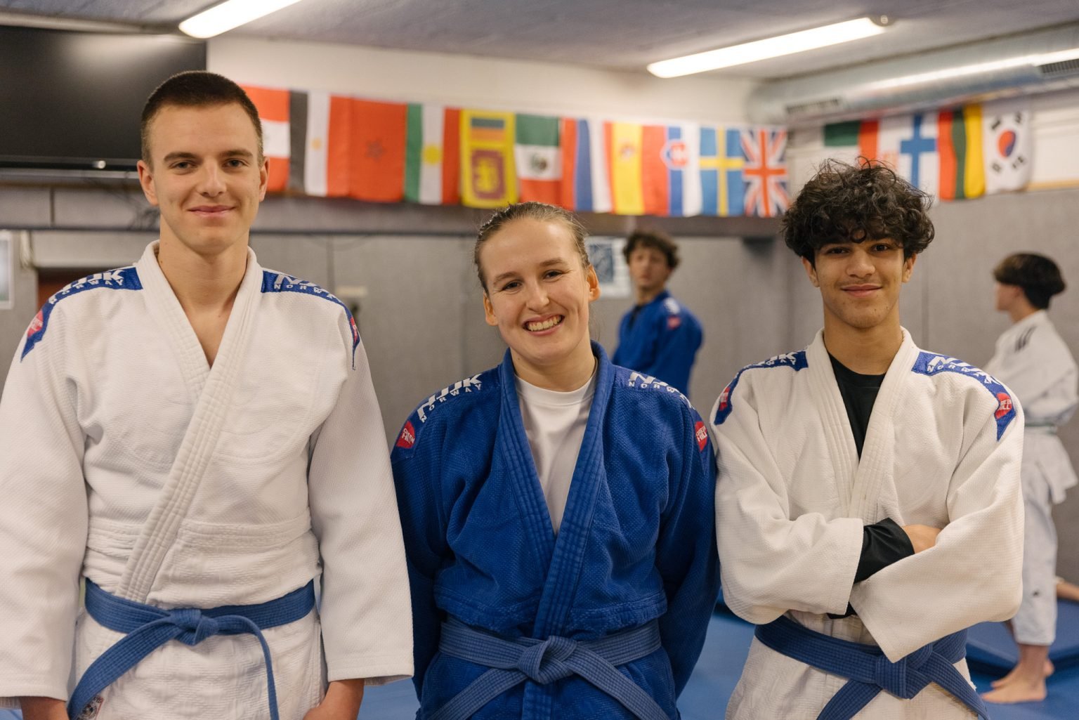 et trenerteam med to gutter iført hvite judodrakter og blått belte, og en jente iført blå judodrakt og blått belte står i en treningssal og smiler inn i kameraet.