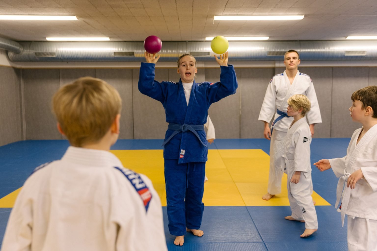 en jente og trener iført blå judodrakt holder i en lilla og en gul softball som hun er på vei til å kaste ut i treningssalen til de unge judoutøverne