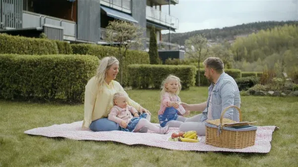 En familie som nyter en piknik på et teppe i gresset, med en kurv og frukt ved siden av, i nærheten av bygårder.