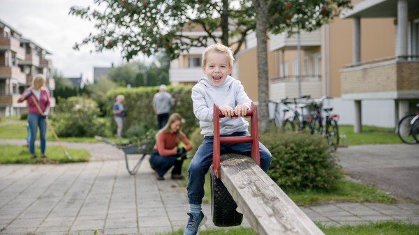 En ung gutt smiler og ler mens han husker i et nabolag, med voksne som gjennomfører dugnad i bakgrunnen.