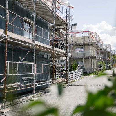 Byggeplass som viser bygninger under oppussing med stillas, omgitt av gjerde og grøntområder på en solrik dag.