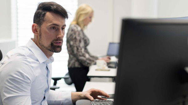 En mann i en hvit skjorte fokuserer på datamaskinen sin på kontoret, og legemliggjør fliden til «Jobb i BORI», mens en kvinne i en mønstret bluse effektivt navigerer den bærbare datamaskinen i bakgrunnen.