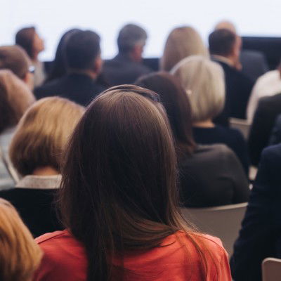 En gruppe mennesker sitter vendt fremover på rader på et innendørs arrangement eller seminar, med ryggen mot kameraet.
