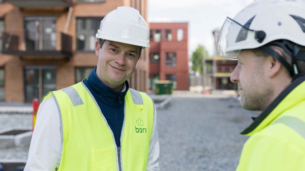To bygningsarbeidere med hvite hjelmer og gule sikkerhetsvester står på en byggeplass og samarbeider med utbyggeren, med moderne boligblokker i bakgrunnen.