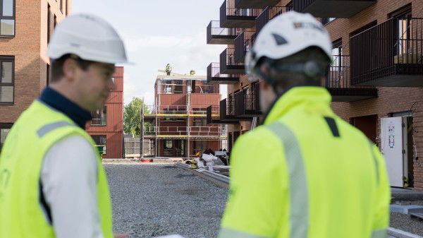 To bygningsarbeidere iført hjelm og refleksvest står foran et leilighetsbygg som er under oppføring i Stifte borettslag.