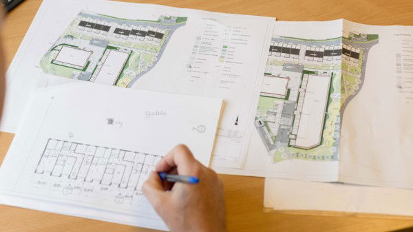 En utbygger tegner en plantegning med penn på papir, omgitt av arkitekttegninger og diagrammer som ligger spredt ut på et trebord.