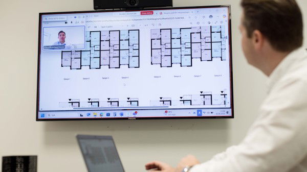 En person på et kontor ser arkitekttegninger fra en utbygger på en stor skjerm under en videosamtale med en annen person, mens en bærbar datamaskin står på skrivebordet.