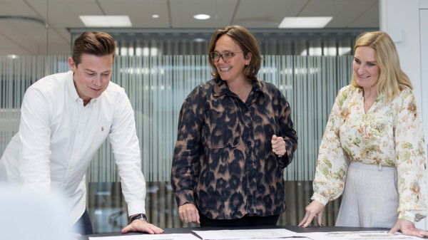Tre personer står rundt et bord og ser på papirer og diskuterer hvordan gjøre eiendom klar for salg i et moderne kontormiljø.