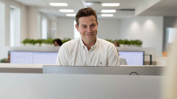 En mann i hvit skjorte sitter ved et skrivebord på et moderne kontor, fokusert på akkvisisjonsoppgaver, med dataskjermer og grønne planter i bakgrunnen.