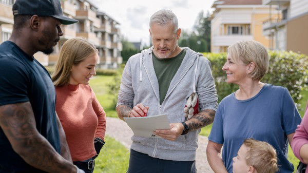 En gruppe mennesker står ute i et boligområde og ser på en mann som holder i en skriveplate og skriver, med bygninger og grøntområder i bakgrunnen.